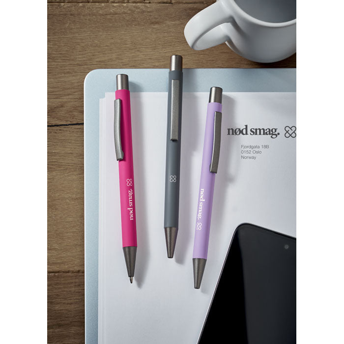 Tres bolígrafos reciclados de aluminio en colores rosa, gris y lila sobre un escritorio, incluyendo el bolígrafo reciclado de aluminio.