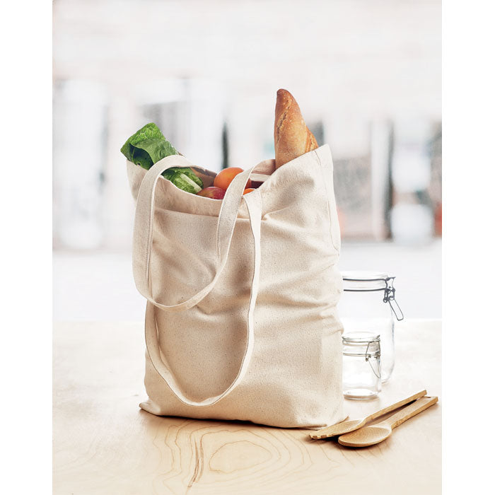 Bolsa de compra RASSA de canvas con fuelle llena de alimentos, ideal para mercados ecológicos. Diseño práctico y reutilizable con asas largas.