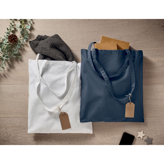 Dos bolsas de algodón ecológico, una blanca y otra azul, sobre una superficie de madera, con etiquetas y contenido visible.
