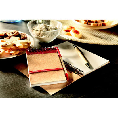 Libreta con espiral en la parte superior y detalles en color rojo, bolígrafo de cartón reciclado y clip, pulsador y punta roja sobre una mesa de cocina