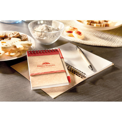 Libreta con espiral en la parte superior y detalles en color rojo personalizada con logotipo en la tapa, bolígrafo de cartón reciclado y clip, pulsador y punta roja sobre una mesa de cocina