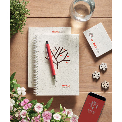 Dos libretas con tapas de papel de semillas personalizadas con un dibujo sobre las que hay un boli rosa y todo ello sobre mesa de madera junto con sets de notas, teléfono móvil y flores