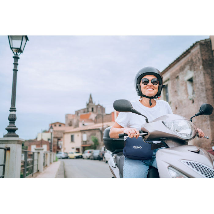 Mujer sonriente con casco y gafas de sol, conduciendo una moto en un paisaje urbano, sosteniendo una riñonera RPET 300D con forro.