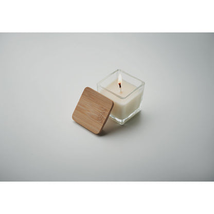 Vela cuadrada blanca con envase de vidrio y tapa de madera a un lado fondo gris