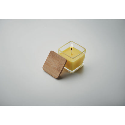 Vela cuadrada amarilla con envase de vidrio y tapa de madera a un lado fondo gris
