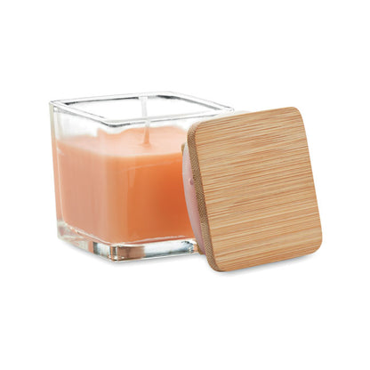 Vela cuadrada naranja con envase de vidrio y tapa de madera a un lado fondo gris