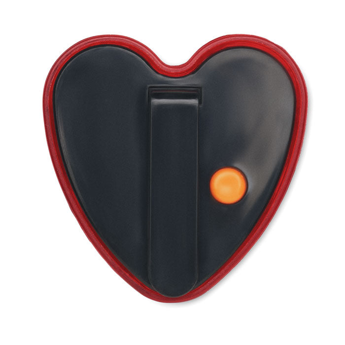 Imagen del reflector en forma de corazón, color negro con borde rojo y botón naranja, con clip trasero para fácil sujeción.