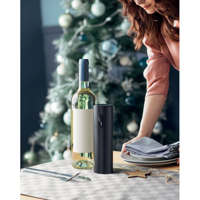 Vista lateral de un abridor eléctrico de botellas negro junto a una botella de vino y utensilios en una mesa decorada.