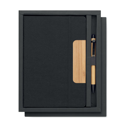 Libreta negra con cierre de goma negra y con pieza de madera en la parte derecha de la portada y bolígrafo de madera de bambú con detalles en color negro. Ambas cosas presentadas en caja negra de cartón