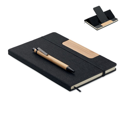 Libreta de poliéster negra tamaño A5 con cierre de goma elástica negra y con bolígrafo de madera de bambú con pulsador, clip y puntera en color negro. La portada tiene una pieza de madera que actua como soporte de teléfono móvil 