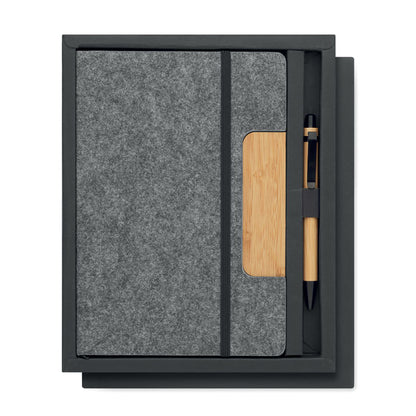 Libreta gris con cierre de goma negra y con pieza de madera en la parte derecha de la portada y bolígrafo de madera de bambú con detalles en color negro. Ambas cosas presentadas en caja negra de cartón