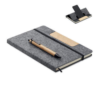 Libreta de poliéster gris tamaño A5 con cierre de goma elástica negra y con bolígrafo de madera de bambú con pulsador, clip y puntera en color negro. La portada tiene una pieza de madera que actua como soporte de teléfono móvil 