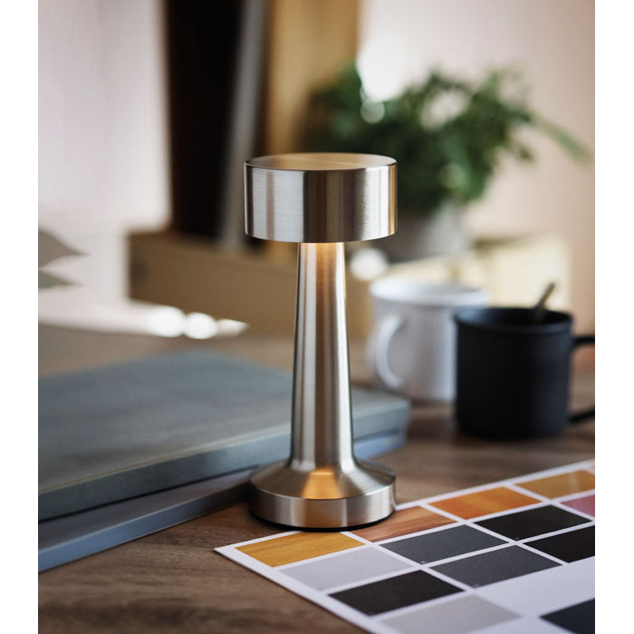 Lámpara de sobremesa de hierro con diseño moderno, acabados en metal, sobre una mesa decorativa junto a paletas de colores y tazas.