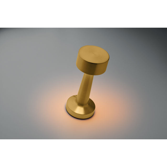 Lámpara de sobremesa de hierro en acabado dorado, con diseño moderno y luz cálida, sobre fondo gris.