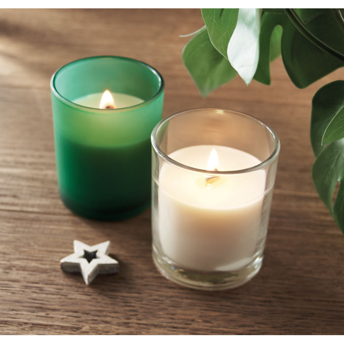 Vista de dos velas encendidas, una verde y otra blanca, sobre una mesa de madera, junto a una planta.