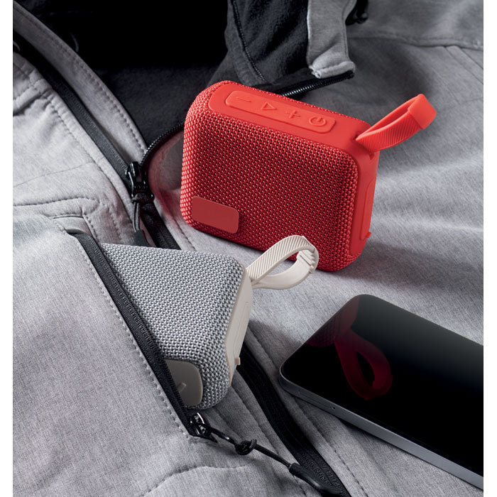 Vista de dos altavoces inalámbricos de 5 W en un bolsillo de chaqueta, uno rojo y otro gris, junto a un teléfono móvil.