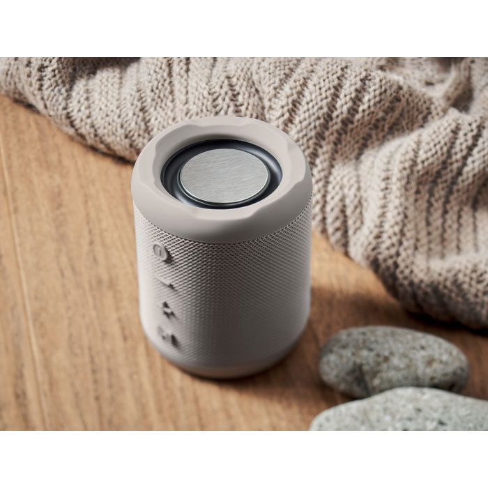 Vista superior de un altavoz inalámbrico ABS de 5 W en color gris, sobre una superficie de madera junto a piedras decorativas.