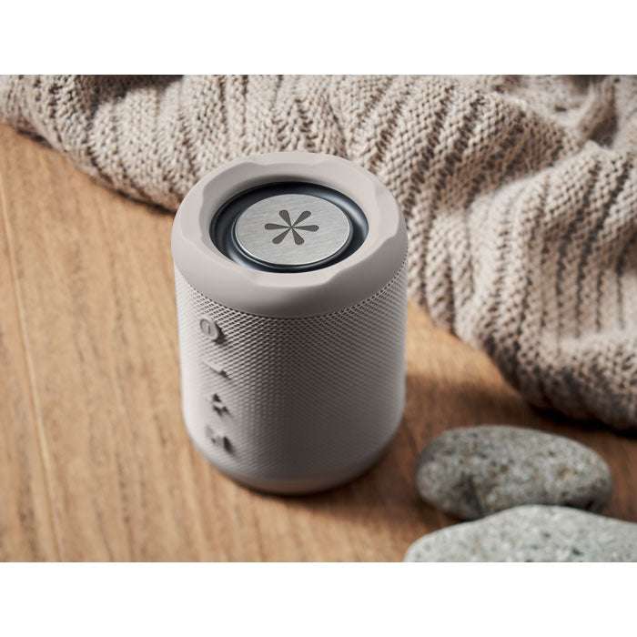 Vista superior de un altavoz inalámbrico ABS de 5 W, color gris, con textura en relieve sobre una mesa de madera.