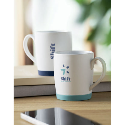 Taza blanca mate con base turquesa en mesa con otra taza al fondo