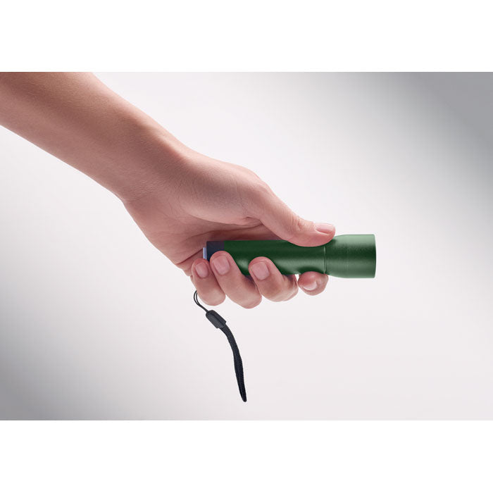 Mano sosteniendo una linterna recargable de aluminio verde con correa, destacando su diseño práctico.