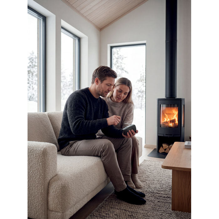 Pareja sentada en un sofá mientras usan un dispositivo móvil junto a una chimenea en un ambiente acogedor.