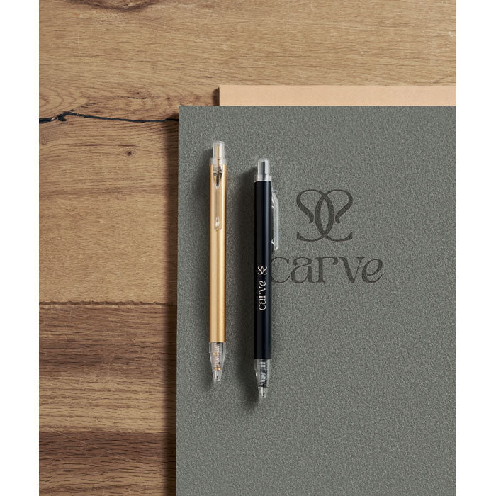 NOSTA - Bolígrafo aluminio reciclado en color dorado y negro sobre una superficie gris con un cuaderno.