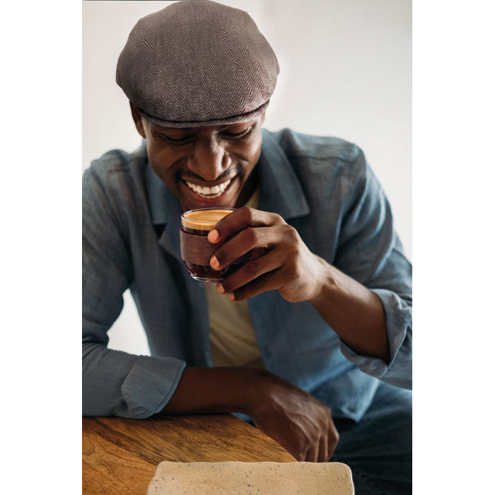 Hombre sonriente disfrutando bebida en vaso del BOGOTA SET - Set vasos café banda de nogal, con gorra y camisa azul.