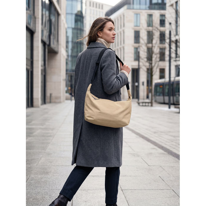 Modelo caminando por la calle con la SLING - Bolsa urbana cruzada de nailon en color beige, usando abrigo gris.