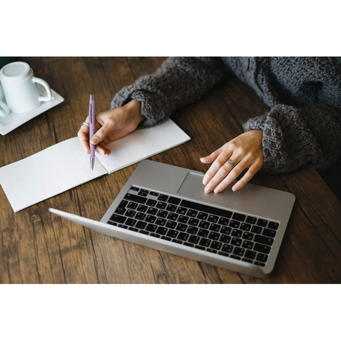 Persona escribiendo en un papel con un bolígrafo reciclado de aluminio sobre una mesa junto a una laptop.