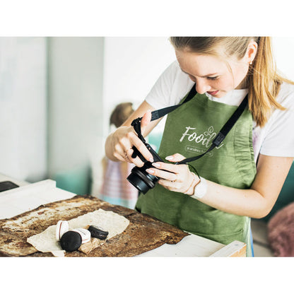 Chica con delantale verde de cáñamo personalizado haciendo una fotografía a varios dulces encima de una mesa
