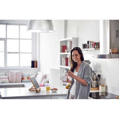 Mujer en cocina modera con móvil y café en las manos y tablet sobre soporte de madera en la mesa 