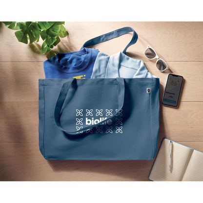 Bolsa de algodón rectangular azul personalizada con logotipo con varias camisetas en su interior y con unas gafas de sol y un teléfono móvil al lado