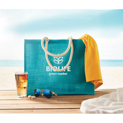 Bolsa de yute turquesa con logo impreso sobre mesa de madera con el mar de fondo