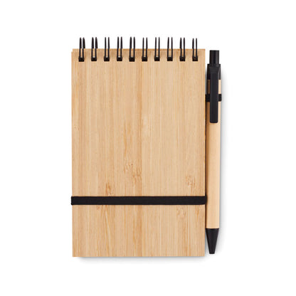 Vista frontal de libreta de bambú con espiral negra en parte superior,, goma de cierre horizontal color negro y bolígrafo de cartón con detalles en color negro
