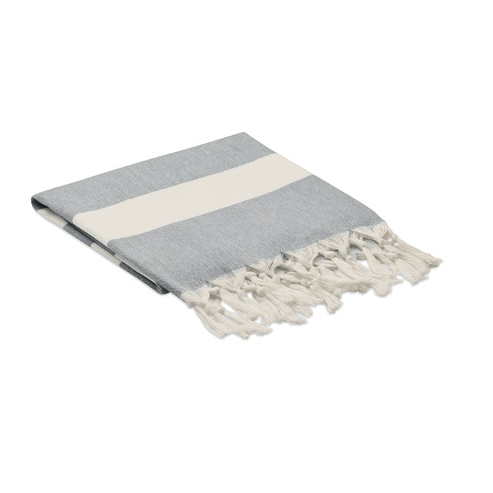 Vista de toalla de playa/pareo en tonos gris y blanco, con flecos decorativos, fabricada en tejidos reciclados.