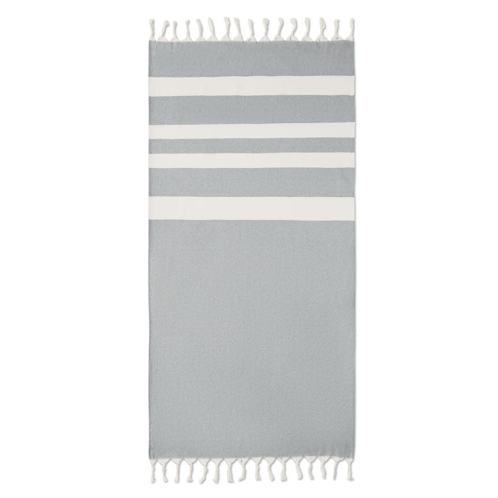 Toalla de playa gris piedra con franjas blancas y flecos, fabricada en tejidos reciclados, ideal para uso en la playa.