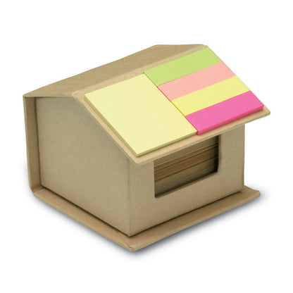 Cajita de cartón en forma de casa con notas adhesivas de colores en el tejado y hojas de papel reciclado en en interior
