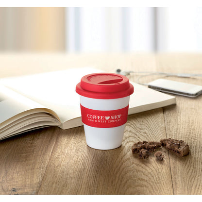 Vaso blanco con talpa y faja de silicona roja personalizado con logotipo sobre mesa de madera al lado de un libro