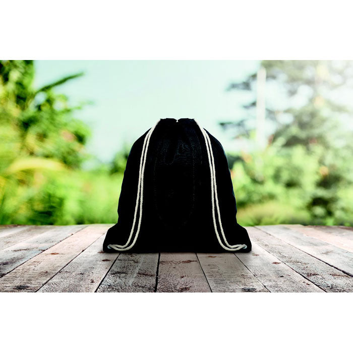 MOIRA - Mochila negra en lona canvas con cordones, fotografiada en exterior sobre superficie de madera, estilo natural y ecológico.