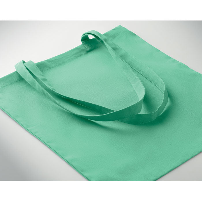 Bolsa de algodón verde con asas largas, material suave y resistente, vista lateral sobre fondo claro