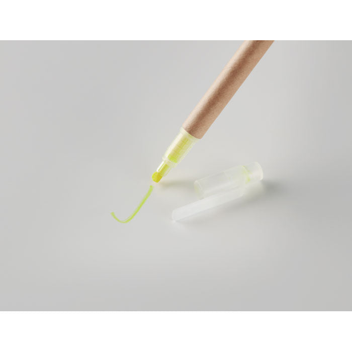 Detalle de un marcador amarillo con cuerpo de cartón y tapa transparente, en primer plano sobre fondo gris.