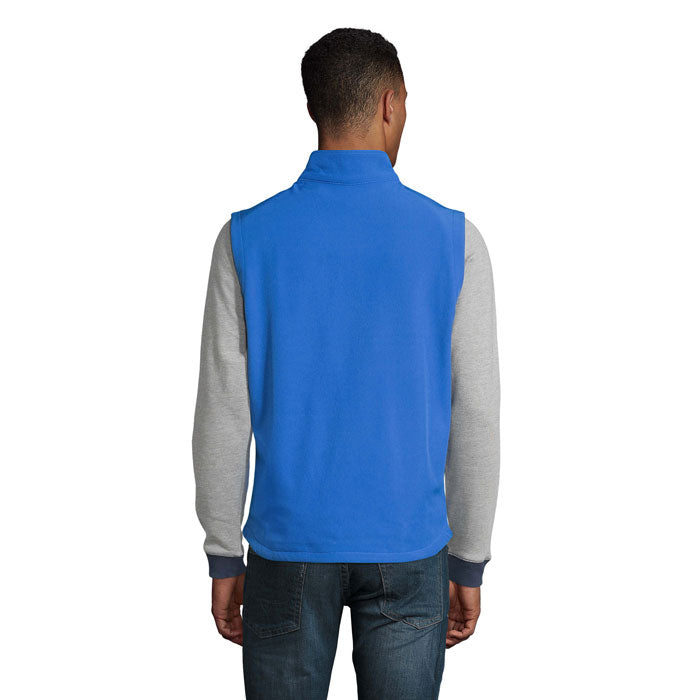 Vista trasera de un chaleco softshell azul para hombre, combinado con mangas grises y pantalones de denim.